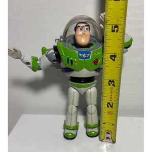 May include: A Buzz Lightyear action figure, featuring a green and white spacesuit with a clear helmet. The figure is approximately 5 inches tall, with arms outstretched. A yellow Stanley measuring tape is next to the figure.