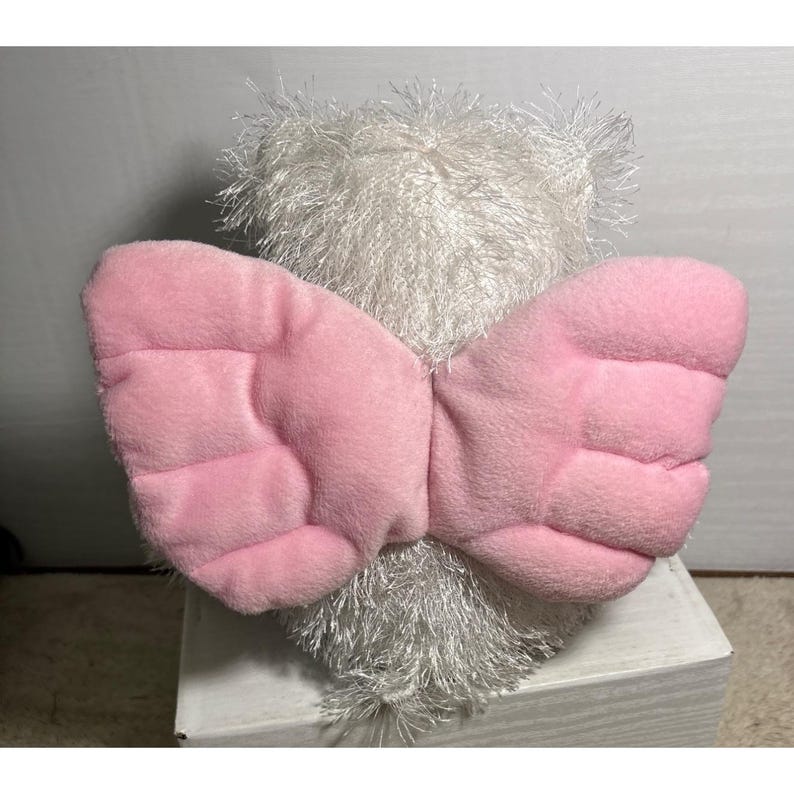 Op de afbeelding: Een witte teddybeer met roze engelen vleugels. De vleugels zijn gemaakt van een zacht, gewatteerd materiaal. De beer heeft een harige, getextureerde vacht. Het speelgoed is ontworpen als een troostrijk item.
