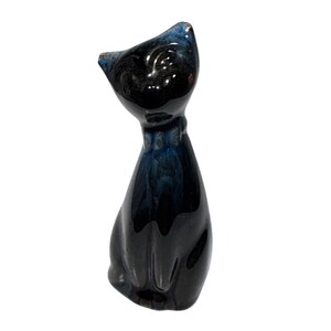 May include: A ceramic cat figurine with a glossy black body and a blue-tinged head and bow tie. The cat is sitting upright, with its head tilted slightly upward. A small red dot is visible on the back of the head. The figurine is a decorative item.