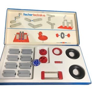 Puede incluir: Una caja abierta de bloques de construcción Fischertechnik 15. La caja contiene una variedad de vigas de plástico gris, engranajes de plástico rojo y ruedas de plástico negro. La caja también incluye una pegatina azul y blanca con las palabras "Fischertechnik" y "Nylon Gear".