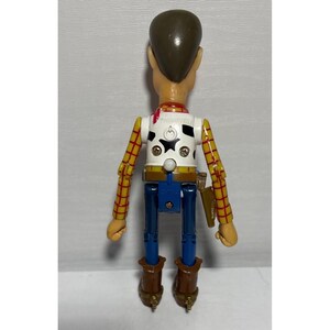 Puede incluir: Una figura de acci&oacute;n de Woody, vista desde atr&aacute;s, con cabeza y pelo casta&ntilde;os, chaleco blanco con estampado de vaca negra y camisa de cuadros amarillos. La figura tiene pantalones azules, botas marrones y una funda marr&oacute;n.