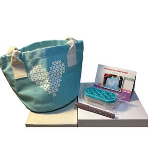 May include: A light blue tote bag with a white cross-stitch heart design. The bag has white handles and sits next to a kit labeled "Small Stitch Love Maker Bucket". Also included is a marker and a container.