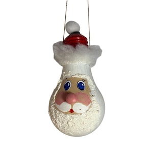 May include: A Christmas ornament shaped like Santa Claus. The ornament has a white bulbous body with a painted face, a red hat, and a white fluffy beard and trim. A gold string is attached for hanging.