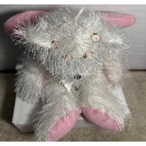 Op de afbeelding: Een witte pluche konijn met roze oren en voeten. Het konijn heeft een pluizige textuur en is versierd met kleine roze rozen rond zijn hoofd. Een witte strik is om zijn nek geknoopt.