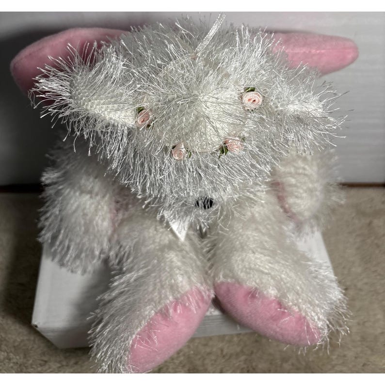 Op de afbeelding: Een witte pluche konijn met roze oren en voeten. Het konijn heeft een pluizige textuur en is versierd met kleine roze rozen rond zijn hoofd. Een witte strik is om zijn nek geknoopt.