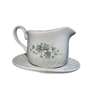 May include: White ceramic gravy boat with a matching saucer. The gravy boat has a curved handle and a spout for easy pouring. It is decorated with a green and gray ivy leaf pattern. The saucer has a ridged edge.