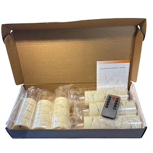 以下が含まれることがあります： 火を使わないキャンドルが入った開いた段ボール箱。キャンドルはアイボリー色で円筒形、ちらつき効果があります。リモコンと取扱説明書も表示されています。取扱説明書のテキストには「火を使わないキャンドルを専門としています」と書かれています。