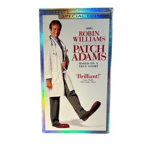 Può includere: Una videocassetta VHS del film Patch Adams, con Robin Williams in camice bianco, stetoscopio e grandi scarpe da clown rosse. La copertina riporta il testo Universal Special Edition, Patch Adams, Based on a True Story e Brilliant!