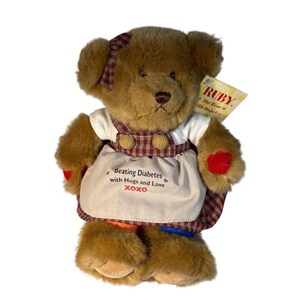 Pode incluir: Um urso de pelúcia castanho vestindo uma blusa branca e um avental xadrez vermelho e branco. O avental tem o texto "Beating Diabetes with Hugs and Love XOXO". O urso tem um laço vermelho e uma etiqueta que diz "RUBY. the bear with Diabetes".