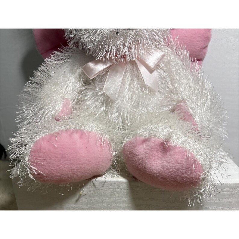 Op de afbeelding: Een zacht wit konijn met roze oren en voeten. Het konijn heeft een pluizig, getextureerd uiterlijk en een delicate roze strik om de nek. Een kinderspeelgoed.