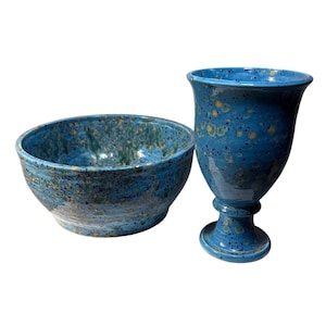 May include: A blue ceramic bowl and goblet set. The bowl is round with a speckled blue and green glaze. The goblet has a pedestal base and a flared rim, also with a speckled blue glaze. The set is handmade.