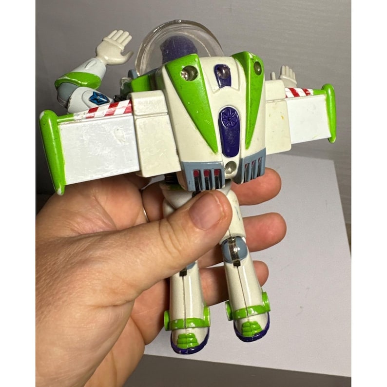 May include: A Buzz Lightyear action figure, featuring a white and green spacesuit design with red and white wing accents. The figure has a clear helmet and is held in a hand, showcasing its articulated limbs and iconic details.