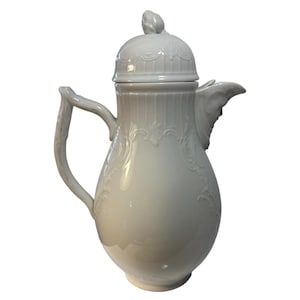 Puede incluir: Cafetera de porcelana blanca con tapa y asa curva. La cafetera presenta diseños florales y de volutas en relieve, y una boquilla con un elemento decorativo. La tapa está rematada con un pequeño adorno en forma de capullo.