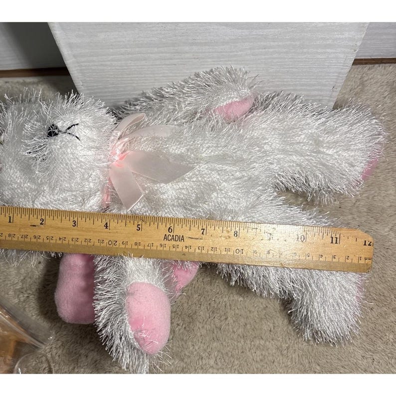 Op de afbeelding: Een pluche wit konijn met roze accenten op de oren en voeten, en een roze strik. Het speelgoed is ongeveer 28 cm lang. Het konijn ligt op een lichtbruin oppervlak.
