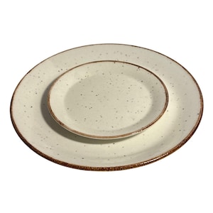 May include: Two off-white ceramic plates, one nested inside the other. The plates have a speckled design with brown rims. The larger plate is round, and the smaller plate is oval-shaped. Dinnerware.