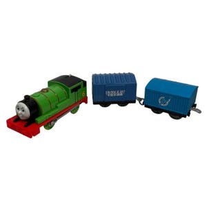 May include: A green toy train with the number 6 on the side, a black roof, and a red front. It is connected to two blue train carriages. One carriage has the text "Frenham Bay" and the other has a globe logo.