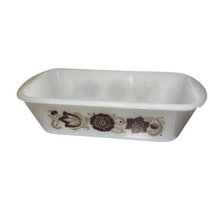 Könnte beinhalten: Eine rechteckige Kastenform aus weißem Milchglas mit einem braunen Blumenmuster an der Seite. Das Design besteht aus stilisierten Blumen und Blättern in verschiedenen Brauntönen, die auf die Außenseite des glatten, opaken Glasgefäßes gedruckt sind.