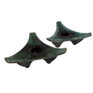 Puede incluir: Dos candelabros de cerámica de color verde azulado oscuro. Cada uno tiene forma triangular, una abertura circular y tres pies redondeados. Los candelabros se muestran sobre un fondo blanco.
