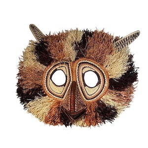 May include: A woven owl mask in shades of brown, tan, and black. The mask has a textured, feathered appearance. The owl's eyes are large, white circles, and the beak is a pointed triangle. The mask has two woven ear-like projections.