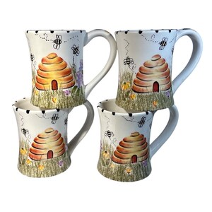 May include: Four white ceramic mugs with a hand-painted bee and beehive design. The mugs feature a beehive, bees, and wildflowers on a grassy background. Each mug has a large handle and a black dotted rim.