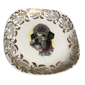 May include: Vintage decorative plate with a poodle portrait. The off-white plate features a gold leaf border and a central illustration of a gray poodle with a red collar against a yellow background. A charming piece for display.