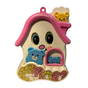 Könnte beinhalten: Ein Vintage-Spielzeughaus aus Kunststoff mit einem lächelnden Gesicht, großen blauen Augen und einem rosa Dach. Zwei blaue Teddybären sind abgebildet, einer in einem Fenster und einer an der Seite. Eine gelbe Maus sitzt auf dem Dach, und die Basis ist mit Blumen verziert.