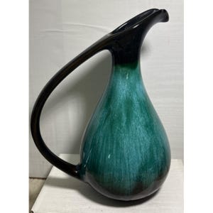 May include: A teal and green ceramic pitcher with a dark brown handle and spout. The pitcher has a teardrop shape and a large, curved handle. The glaze creates a marbled effect on the surface of the pitcher.