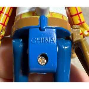 Puede incluir: Primer plano de la parte trasera de una figura de juguete, con pantalones azules, cintur&oacute;n marr&oacute;n y camisa amarilla con detalles rojos. La palabra "CHINA" est&aacute; grabada en blanco sobre el pl&aacute;stico azul. Se ve un tornillo peque&ntilde;o.