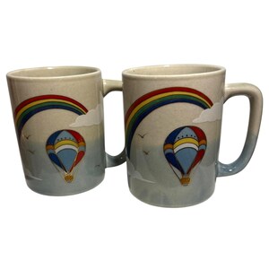 May include: Two ceramic mugs with a light blue gradient. Each mug features a rainbow, clouds, and a hot air balloon design in red, blue, yellow, and white. The mugs have a curved handle.
