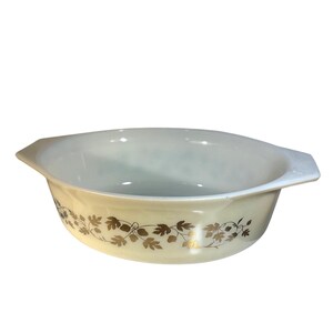 May include: An oval-shaped white glass casserole dish featuring integrated side handles and a decorative band of metallic gold leaves and vines wrapping around the exterior. The interior is smooth and white, with a glossy finish throughout.