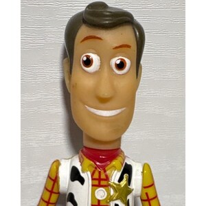 Puede incluir: Primer plano de una figura de acci&oacute;n de Woody de la franquicia Toy Story. La figura tiene una cara bronceada, cabello casta&ntilde;o y viste un atuendo de sheriff con una insignia de estrella dorada. Los ojos de la figura son marrones.