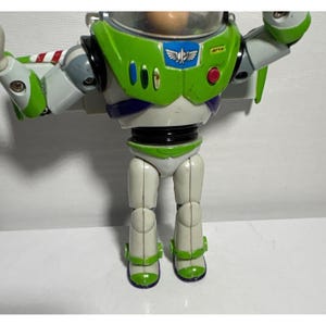 May include: A Buzz Lightyear action figure, featuring a green and white spacesuit design. The figure has a clear helmet, blue and white accents, and the iconic "Star Command" logo. The toy's arms are outstretched, and it stands on a white surface.