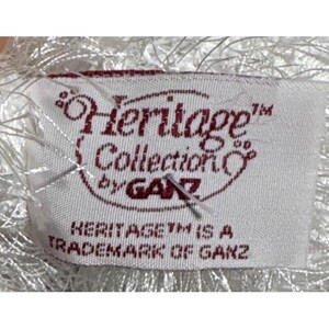 Op de afbeelding: Een wit stoffen label met de tekst "Heritage Collection by GANZ" in kastanjebruine letters. Het label bevat ook de tekst "HERITAGE TM IS A TRADEMARK OF GANZ". Het label is bevestigd aan een wit, pluizig materiaal.