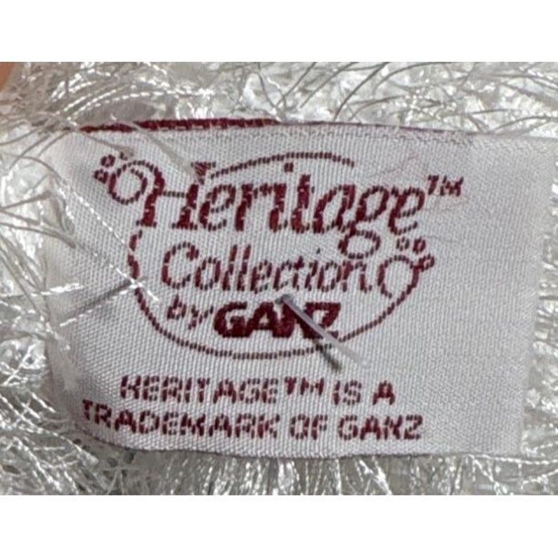 Op de afbeelding: Een wit stoffen label met de tekst "Heritage Collection by GANZ" in kastanjebruine letters. Het label bevat ook de tekst "HERITAGE TM IS A TRADEMARK OF GANZ". Het label is bevestigd aan een wit, pluizig materiaal.