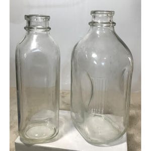 Puede incluir: Dos botellas de vidrio transparente. Una es rectangular con cuello estrecho, la otra tiene forma cuadrada redondeada con cuello más ancho. Ambas botellas están vacías y parecen ser vintage o antiguas.