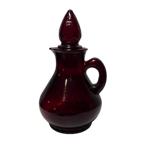 Puede incluir: Una aceitera de vidrio rojo rubí con asa y tapón puntiagudo. La aceitera tiene una base redondeada y un pequeño pico. El asa tiene forma de lazo. El tapón tiene forma de lágrima.