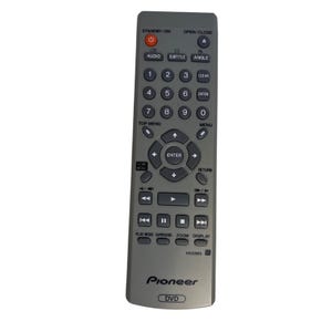 Könnte beinhalten: Eine graue Pioneer DVD-Fernbedienung mit verschiedenen Tasten, darunter Ein/Aus, Audio, Untertitel und Zifferntasten. Die Fernbedienung hat ein Steuerkreuz und ein "DVD"-Etikett unten. Modell VXX2913.