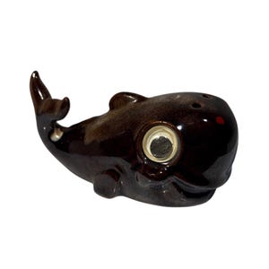 May include: A brown ceramic whale ornament with a shiny glaze. The whale features a large, circular eye with a silver surround and a red spot on its head. It has two holes on top, possibly for salt or pepper.