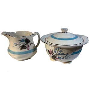 May include: Cream-colored ceramic creamer and covered bowl set. The set features a blue stripe and floral design with black, blue, and pink accents. The creamer has a handle and spout, while the bowl has a lid.