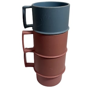 Könnte beinhalten: Drei gestapelte Becher. Der obere Becher ist ein gedämpftes Blau, und die beiden unteren Becher sind rotbraun. Jeder Becher hat einen quadratischen Griff. Die Becher scheinen aus einem strukturierten Kunststoffmaterial zu bestehen.
