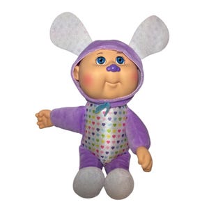 Puede incluir: Muñeca Cabbage Patch Kids con un disfraz de conejito morado. La muñeca tiene ojos azules, mejillas rosadas y una nariz morada. El mono tiene un estampado de corazones coloridos. Las orejas de conejo son blancas con el interior lila claro.