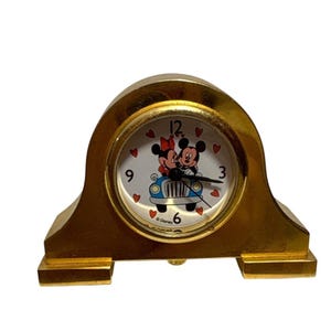 May include: A gold-colored, vintage Mickey and Minnie Mouse clock. The clock face features Mickey and Minnie in a car, surrounded by hearts. The clock has a classic arched shape and a small, round clock face.