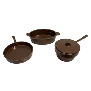 May include: Three brown toy kitchen items: a frying pan with a handle, an oval baking dish, and a pot with a lid. The items are made of a solid, dark brown material and are arranged on a white background.
