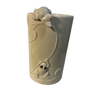Puede incluir: Un vaso cilíndrico de cerámica de color crema con un diseño en relieve de dos ranas trepando por una enredadera. Una rana está situada cerca del borde superior, mientras que la otra está más abajo, ambas integradas en la superficie lisa.