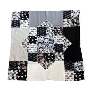May include: A patchwork quilt with a cat theme. The quilt is made up of squares of black, white, and grey fabrics, some with cat faces, cat illustrations, and cat photos. The quilt has a diamond-shaped central design.
