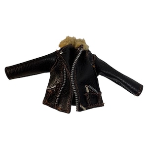 Könnte beinhalten: Eine Miniatur-Lederjacke in Braun mit einem Kunstfellkragen. Die Jacke hat einen Reißverschluss und zwei kleine Taschen. Die Ärmel sind ausgebreitet und die Jacke scheint neu zu sein.
