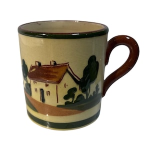 May include: Cream ceramic mug with a brown handle and rim. The mug features a hand-painted landscape scene with a house, trees, and a path. The mug has a dark green and brown colour scheme.