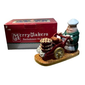 May include: A ceramic figurine of a baker pushing a cart filled with pies and bread. The baker wears a white hat and apron with a green robe. The figurine is on a textured base, with a red box labelled "Merry Makers" in the background.