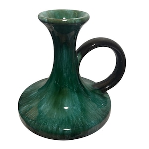 May include: A ceramic candlestick holder with a dark green and black glaze. The holder has a wide base, a slender neck, and a loop handle. The interior is a lighter green.