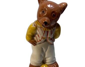 May include: A ceramic bear figurine with a brown head and paws, wearing a yellow shirt and white pants. The vest has red stripes. The bear is standing with its hands behind its back, and its head is tilted.